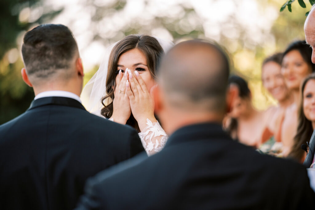 A bride covers her mouth with her hands, appearing emotional during an outdoor wedding ceremony. Groomsmen and bridesmaids are visible in the background.