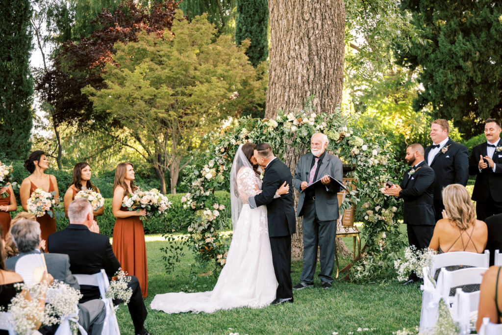 A bride and groom kiss at an outdoor wedding ceremony, surrounded by their bridal party and guests, in front of a floral arch and large tree.
