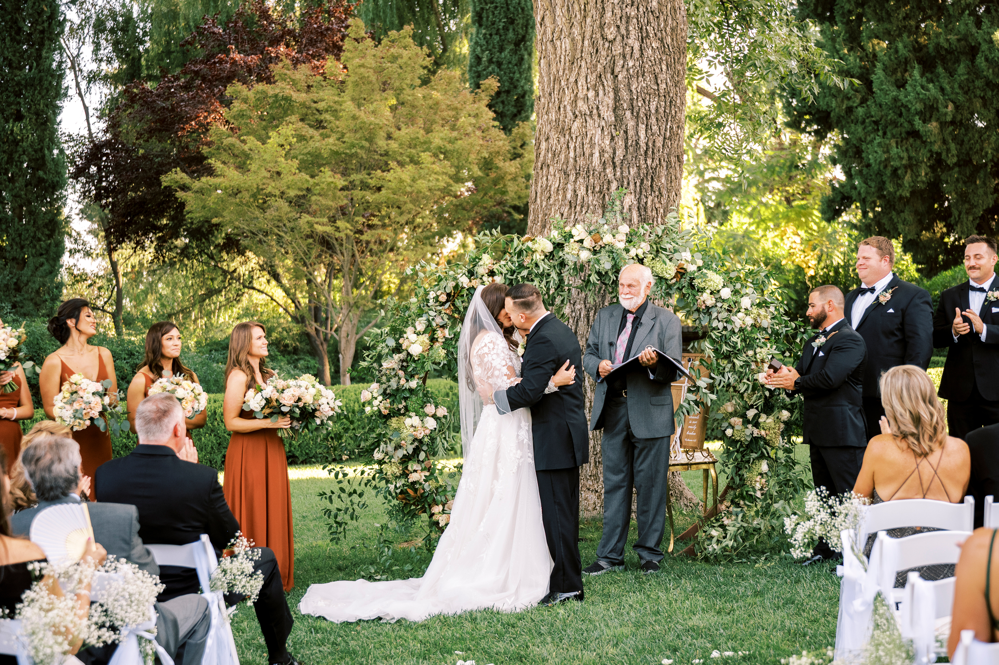 A bride and groom kiss at an outdoor wedding ceremony, surrounded by their bridal party and guests, in front of a floral arch and large tree.