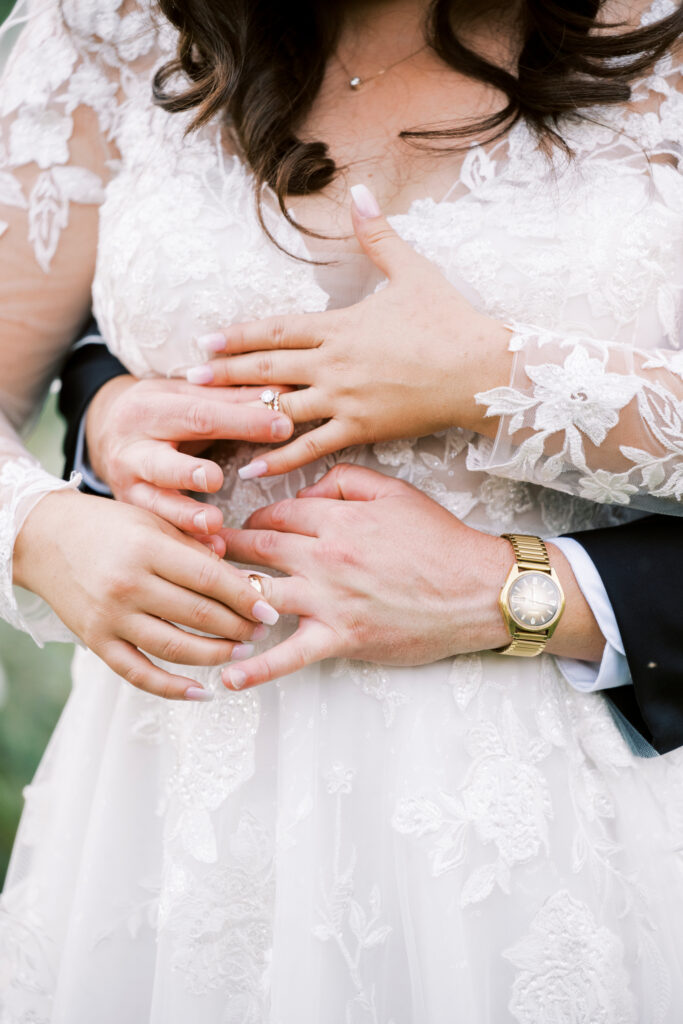 A close-up of a couple's hands, with the bride in a lace dress showing her ring, and the groom wearing a gold watch, embracing from behind.