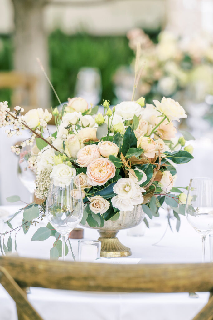 A floral arrangement with white and pale peach roses, greenery, and eucalyptus in a gold vase, set on a white table with wine glasses and a wooden chair.