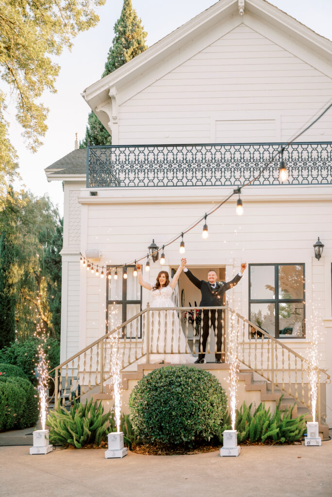 A bride and groom stand on outdoor steps of a white house, holding hands with arms raised, as sparklers shoot up around them. String lights are overhead.