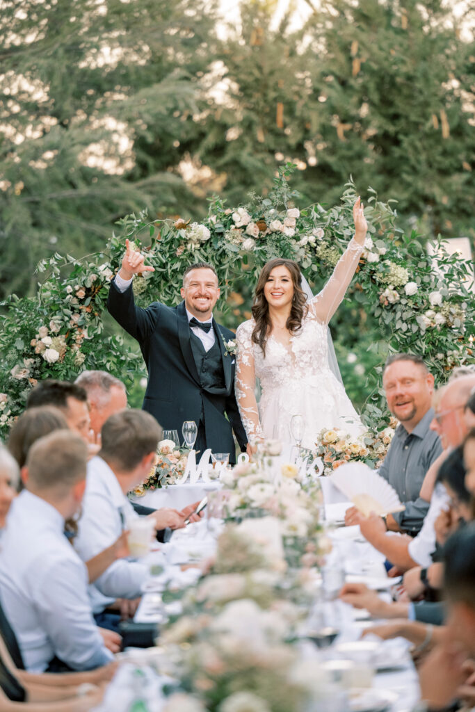 A bride and groom stand smiling and waving at the head of a long outdoor reception table, surrounded by guests and floral decorations.