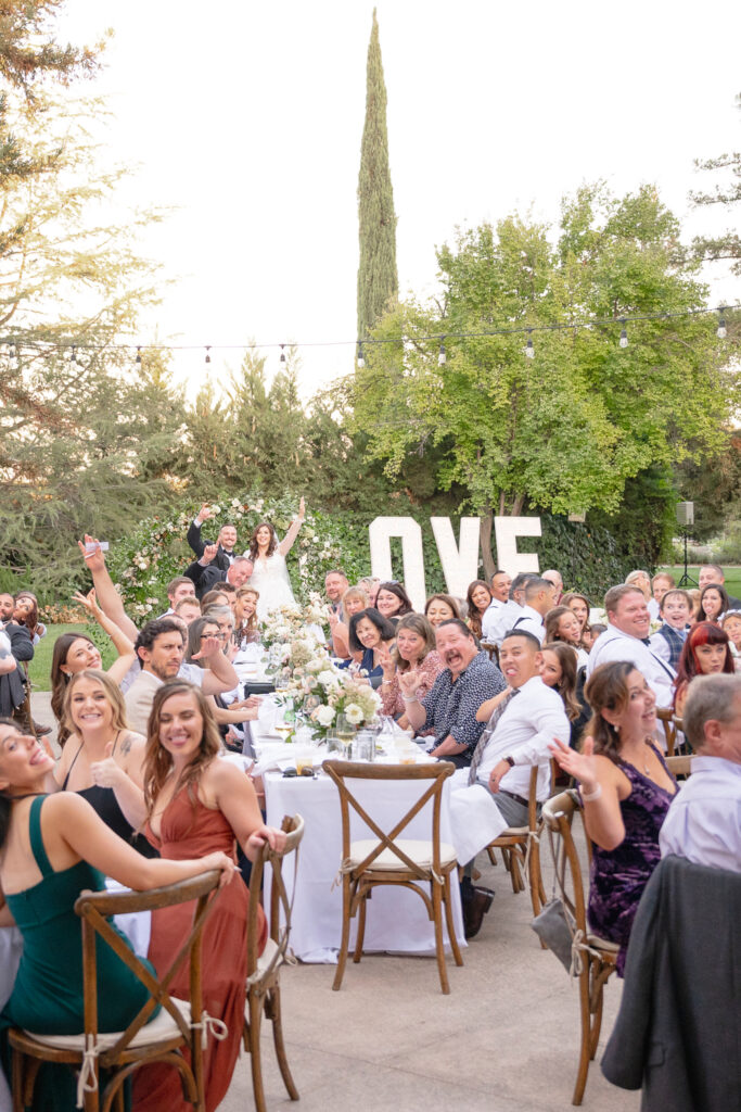 A large group of people seated at outdoor tables, smiling and looking toward the camera, with large "LOVE" letters and greenery in the background.