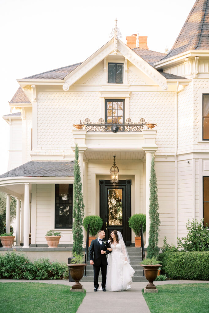 A bride and groom walk together outside a large, elegant Victorian-style house with potted plants and manicured bushes.