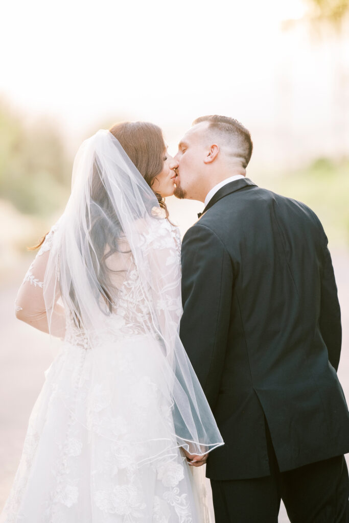 A bride in a white lace dress and veil kisses a groom in a black tuxedo outdoors while holding hands.