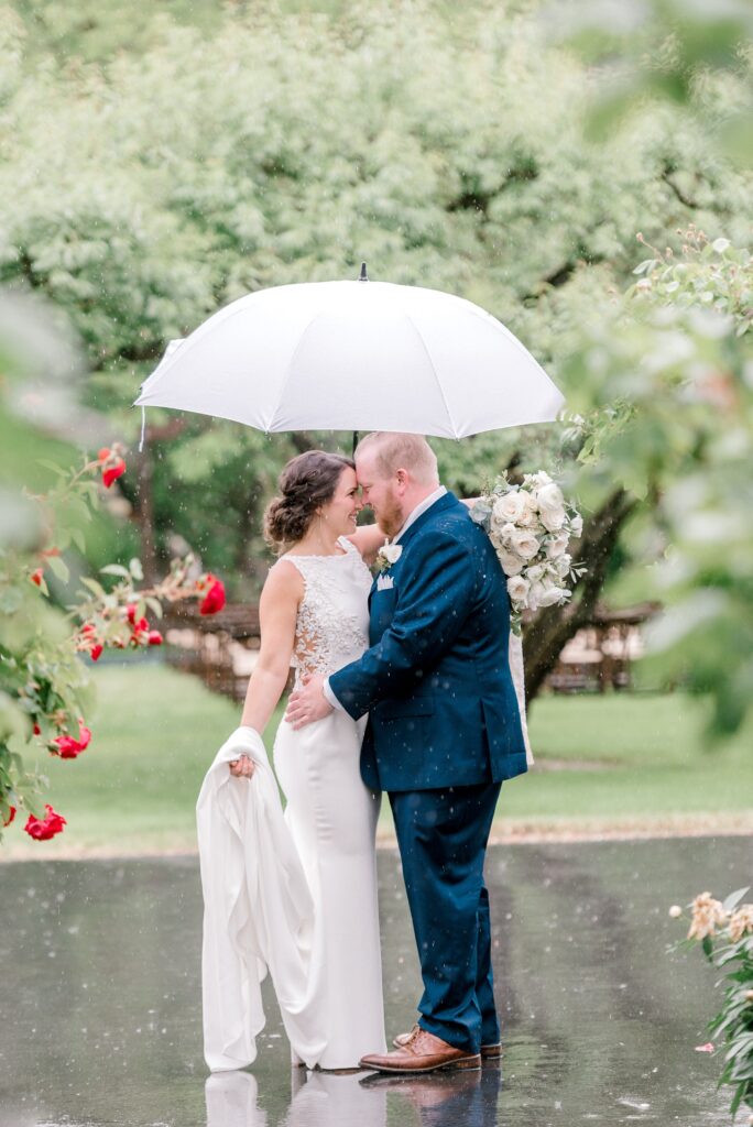 A bride and groom stand close together under a white umbrella in the rain, surrounded by greenery and red flowers. The bride holds her dress, and the groom holds a bouquet.