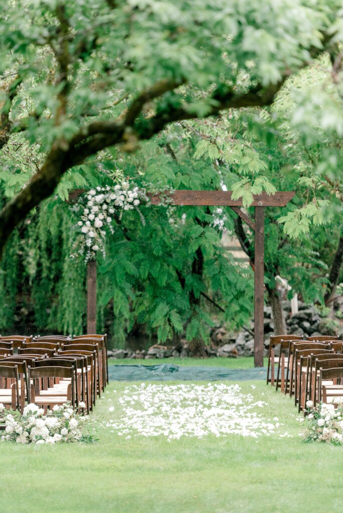 An outdoor wedding ceremony setup with rows of wooden chairs, a wooden arch decorated with white flowers, and flower petals scattered along the grass aisle.