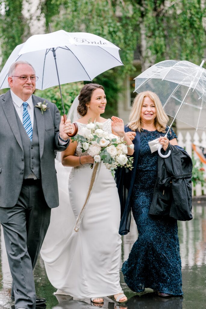 A bride holding a bouquet walks between two people under umbrellas on a wet day, with a man in a suit and a woman in a navy dress.