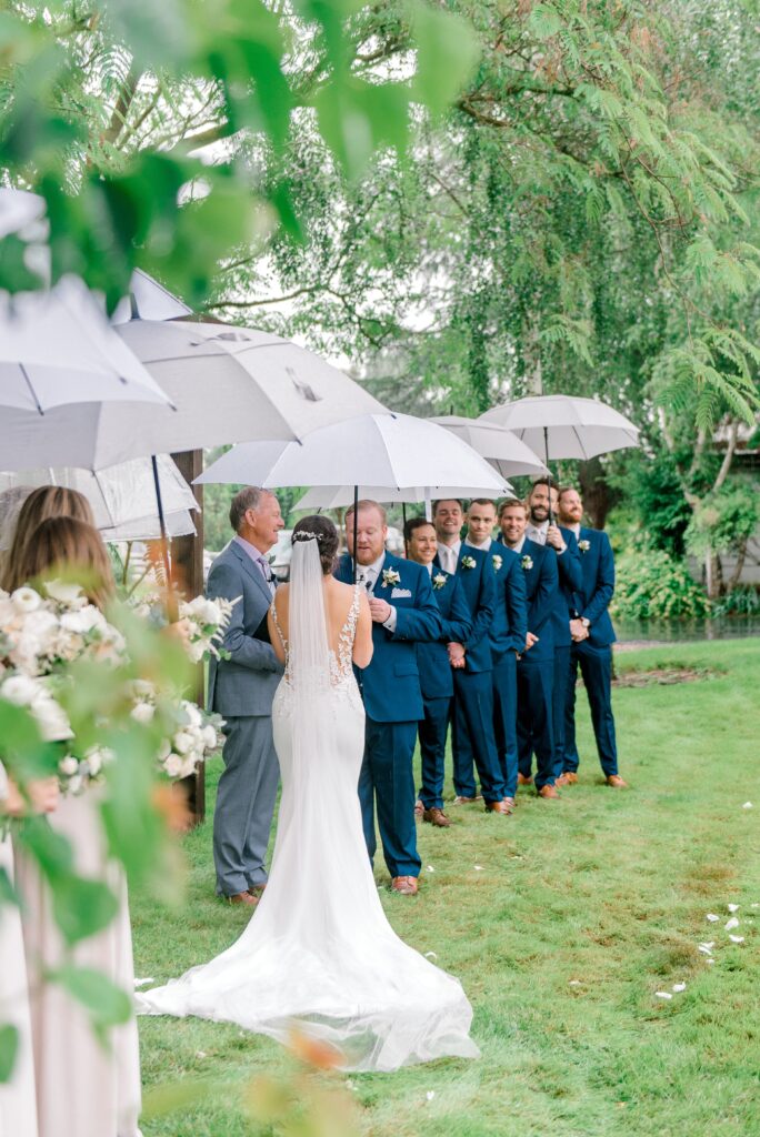 A bride and groom stand facing each other during an outdoor wedding ceremony, with the groom and groomsmen holding umbrellas in the rain.