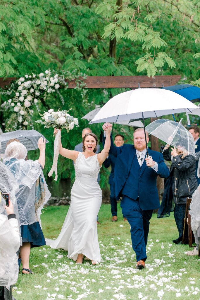 A bride and groom walk outdoors under umbrellas, holding hands and smiling, surrounded by guests with umbrellas and rain ponchos at a wedding ceremony.