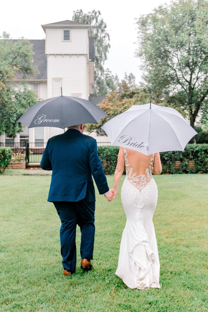 A bride and groom walk hand in hand on grass, holding umbrellas labeled "Bride" and "Groom," with a white building and trees in the background.