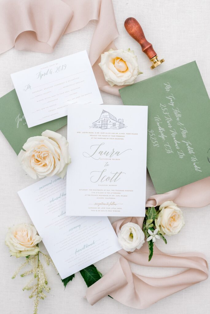 A flat lay of wedding invitations, an olive green envelope with calligraphy, cream-colored roses, and a wax seal stamp on a light fabric background.