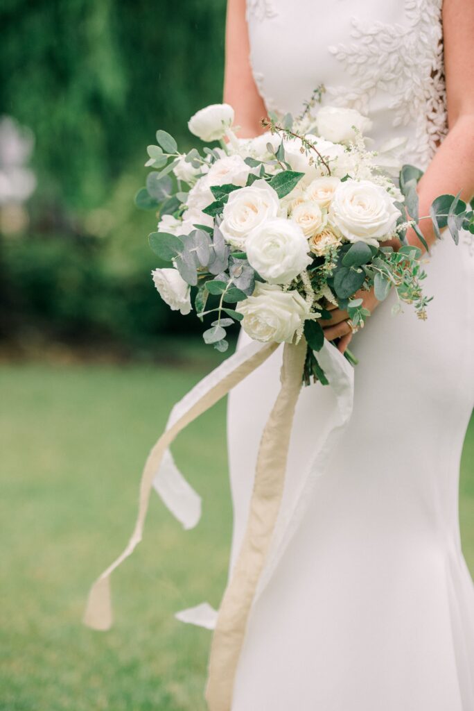 A bride in a white dress holds a bouquet of white and cream flowers with greenery and long ribbon streamers, standing on grass.