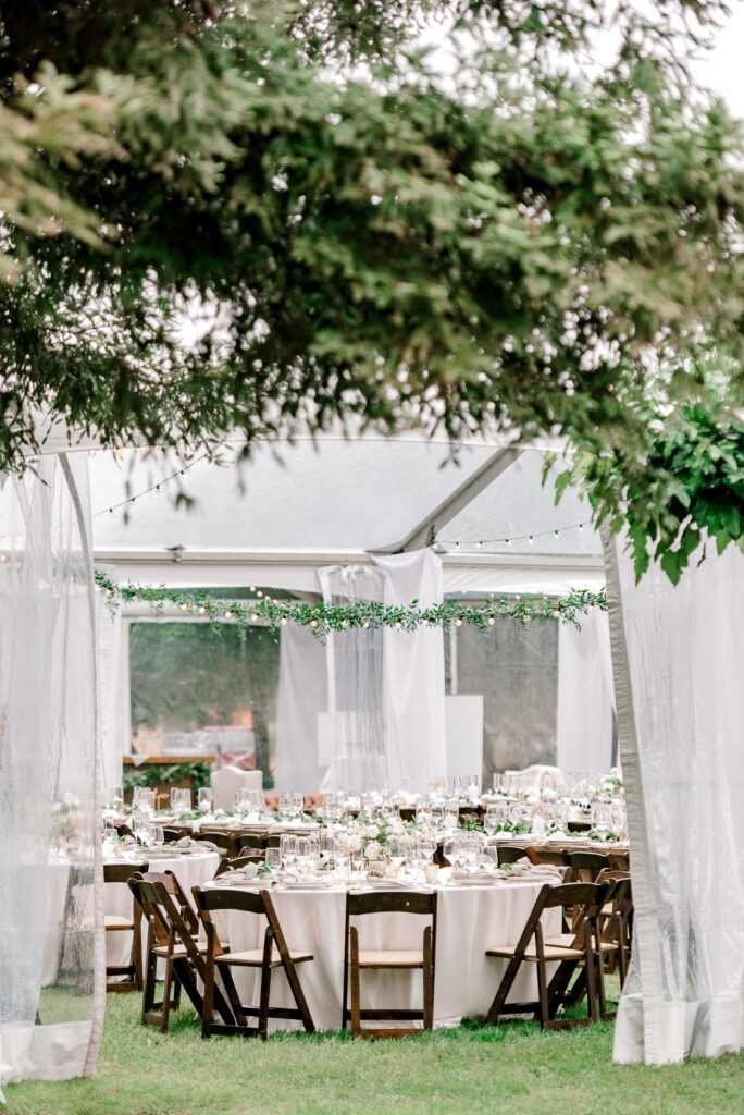 Outdoor event setup with round tables, white tablecloths, glassware, and wooden chairs under a clear tent, surrounded by greenery.