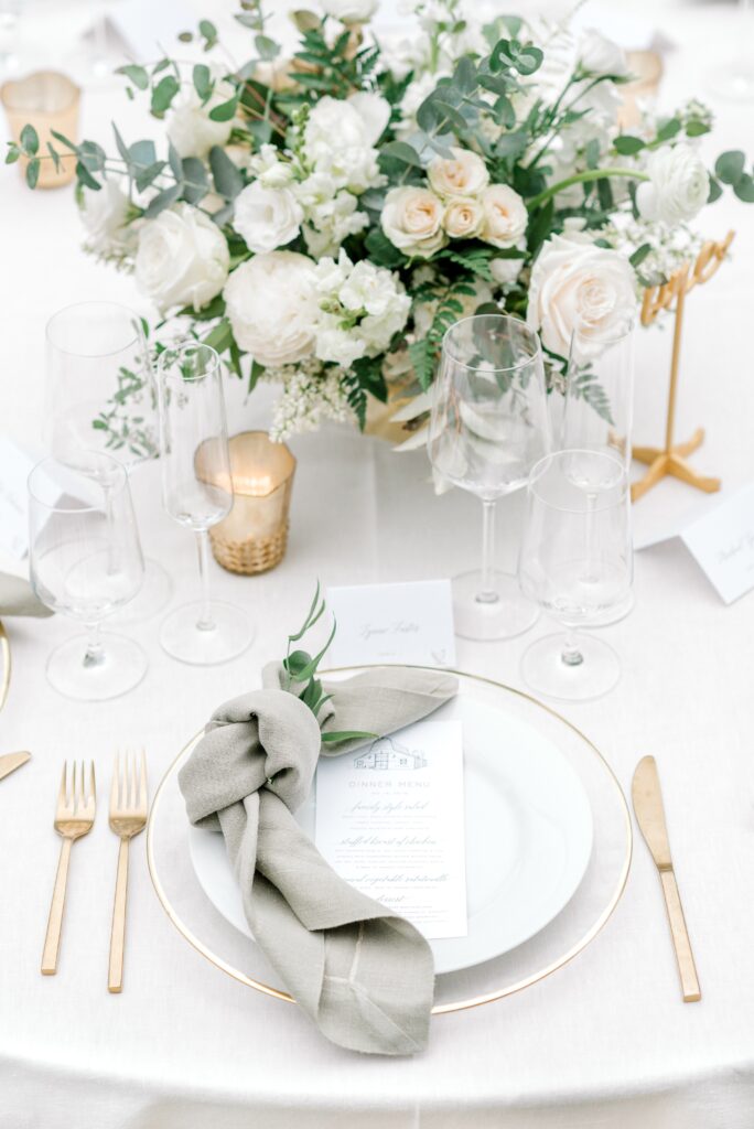 Elegant table setting with gold cutlery, a white plate topped with a menu and knotted napkin, wine glasses, and a floral centerpiece with white and blush flowers.