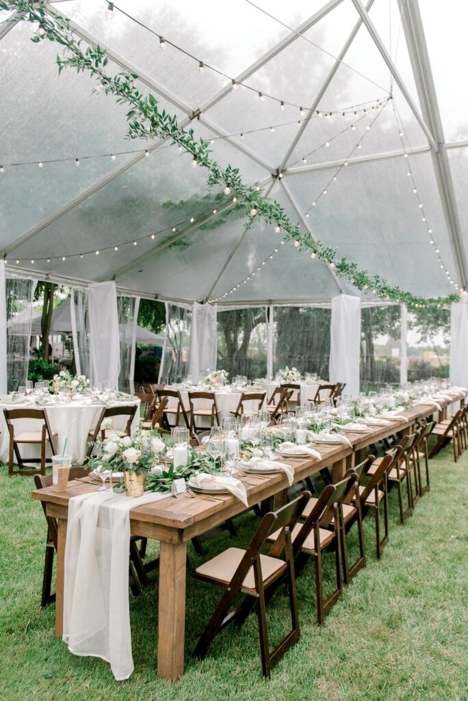 Outdoor wedding reception setup under a clear tent with string lights, long wooden tables, draped white linens, floral centerpieces, and wooden chairs on green grass.