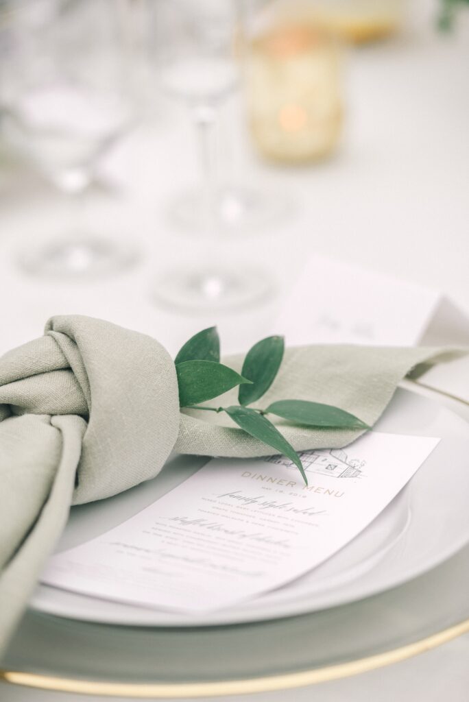 A close-up of a formal dinner place setting with a white plate, a printed menu, a sage green napkin, and a sprig of greenery, set on a white tablecloth.