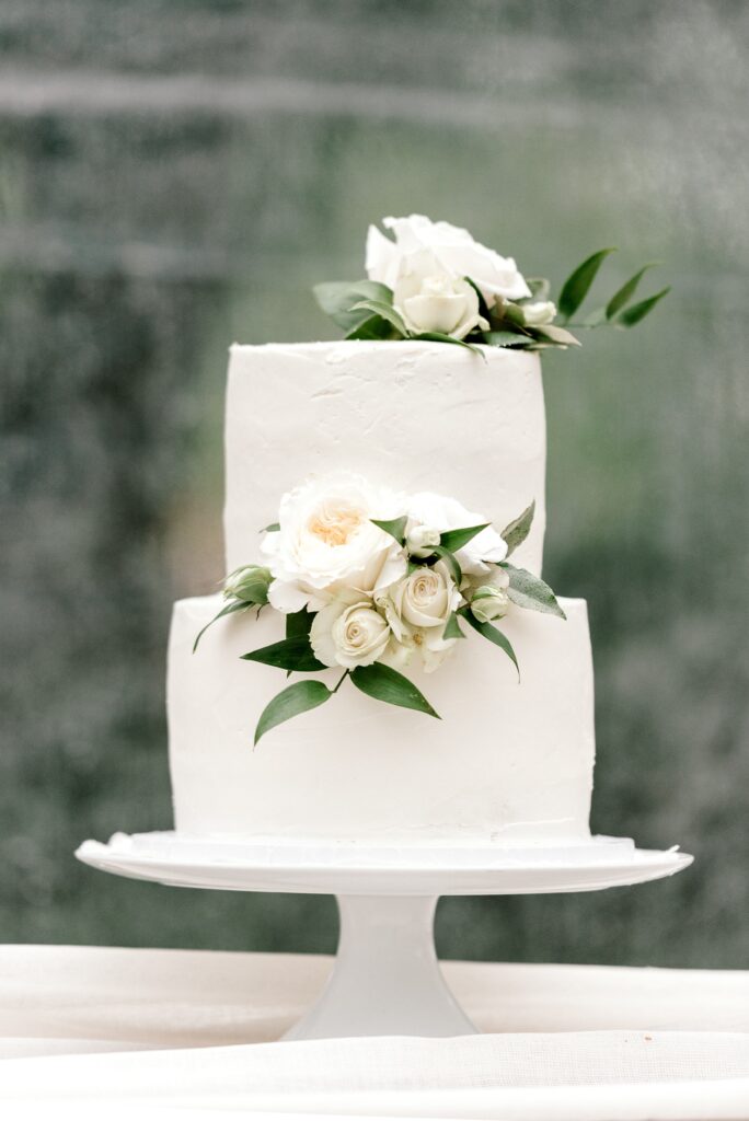 Two-tiered white cake decorated with white roses and green leaves, displayed on a white cake stand against a blurred background.