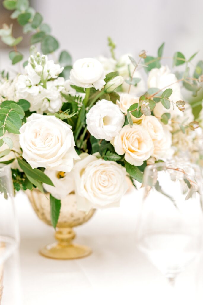 A gold vase holds a floral arrangement of white and pale cream roses, ranunculus, greenery, and eucalyptus on a light-colored table.