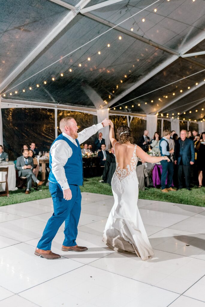 A bride and groom dance on a white floor under a clear tent with string lights, while guests watch from their tables in the background.