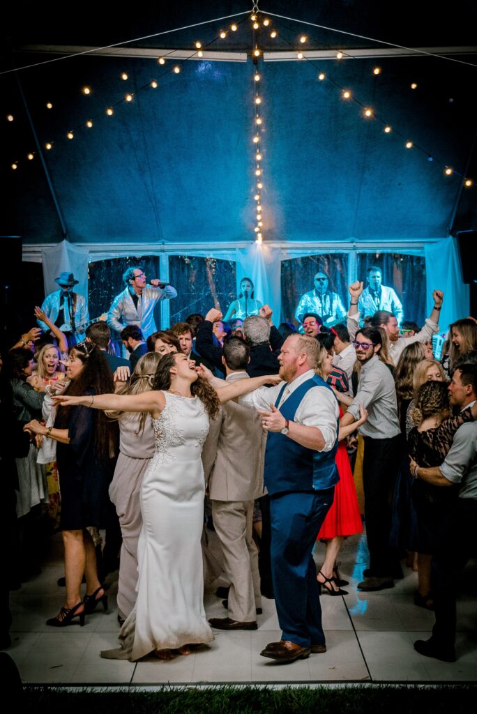 A large group of people dance closely together under string lights at a wedding reception, with a bride and groom celebrating in the center.
