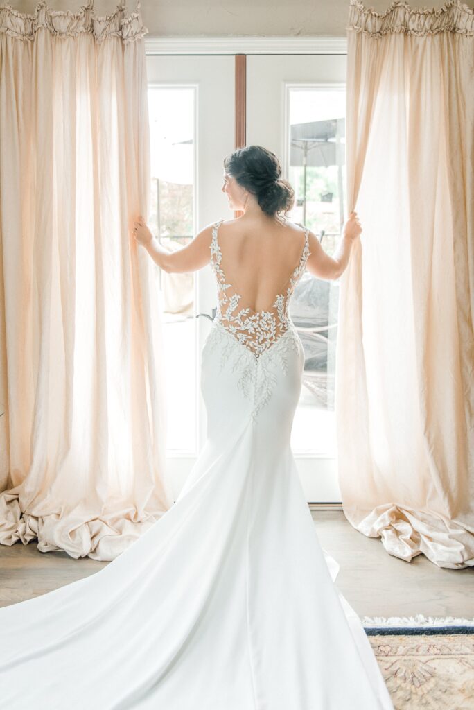 A bride in a white lace-back wedding gown stands facing a window, holding open light curtains, with natural light illuminating the room.