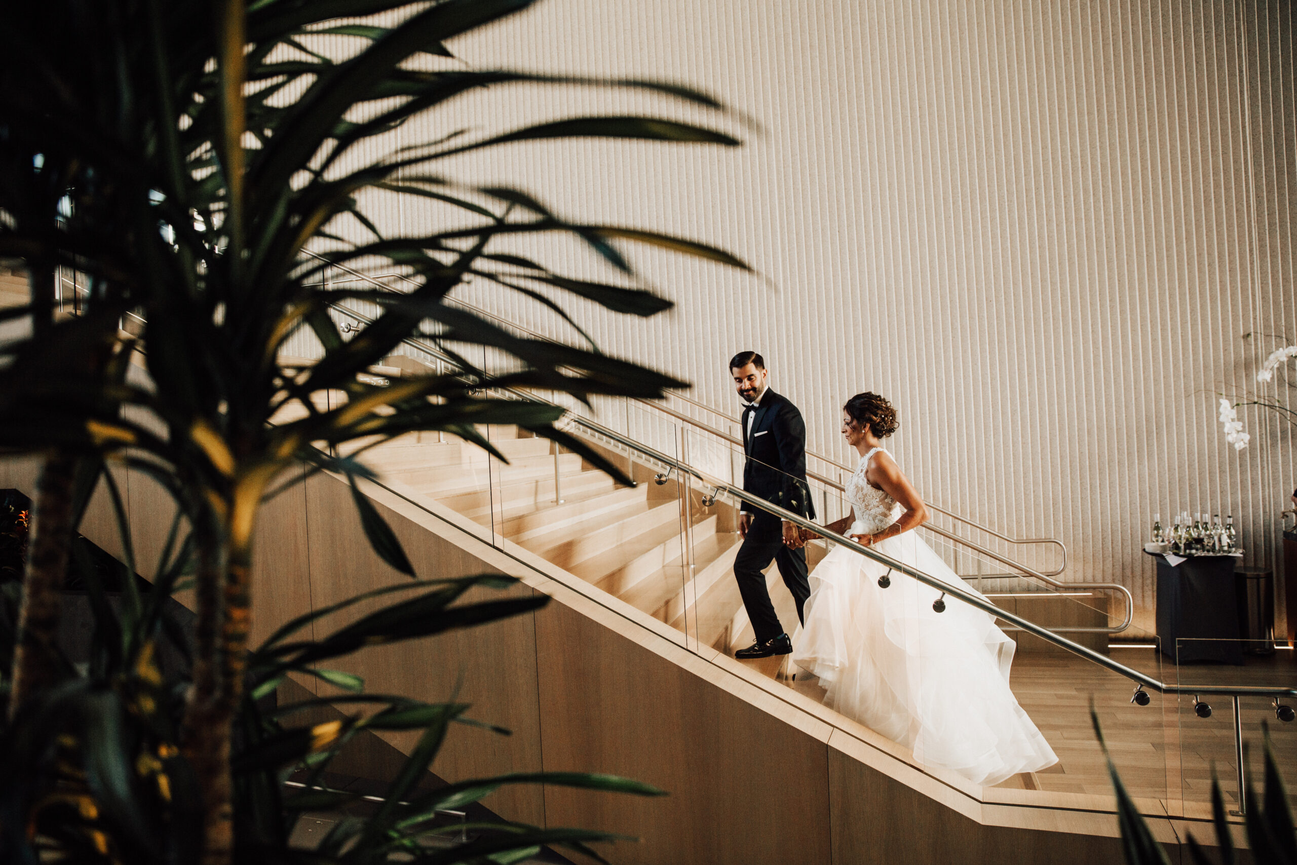 A bride and groom walk up a wooden staircase, with large plants in the foreground and decorative vertical paneling on the wall.