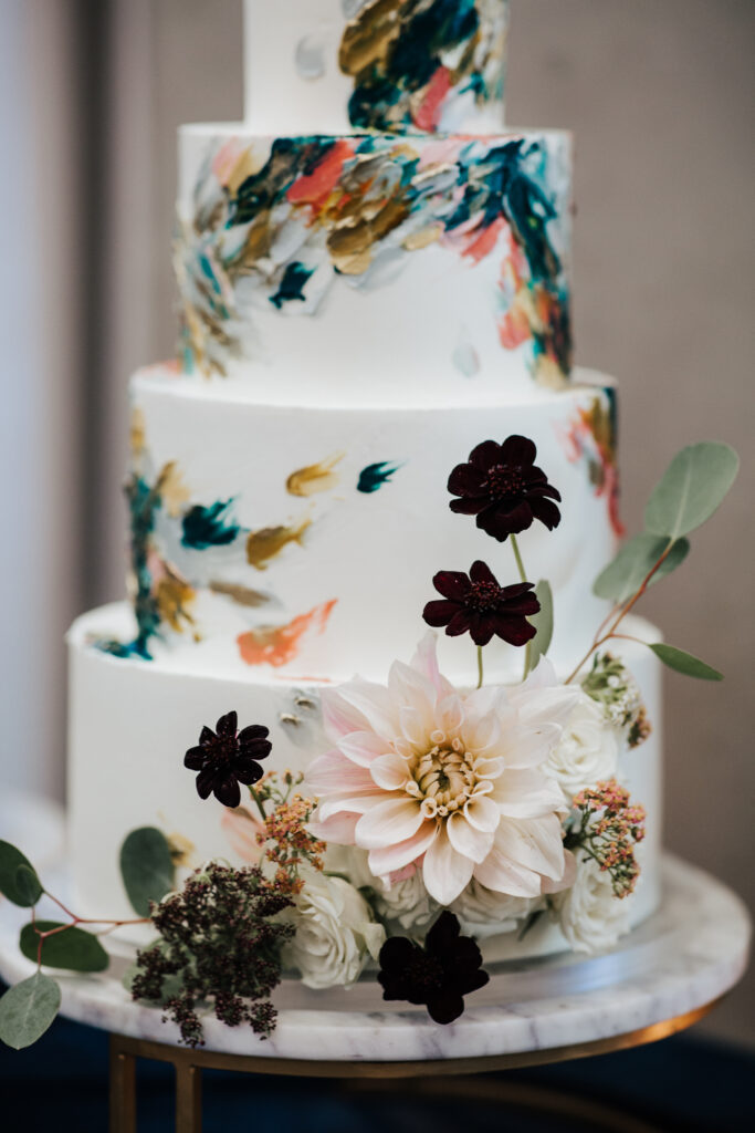 Three-tiered cake with colorful brushstroke accents, adorned with a large pink dahlia, dark flowers, greenery, and small white roses, set on a marble base.