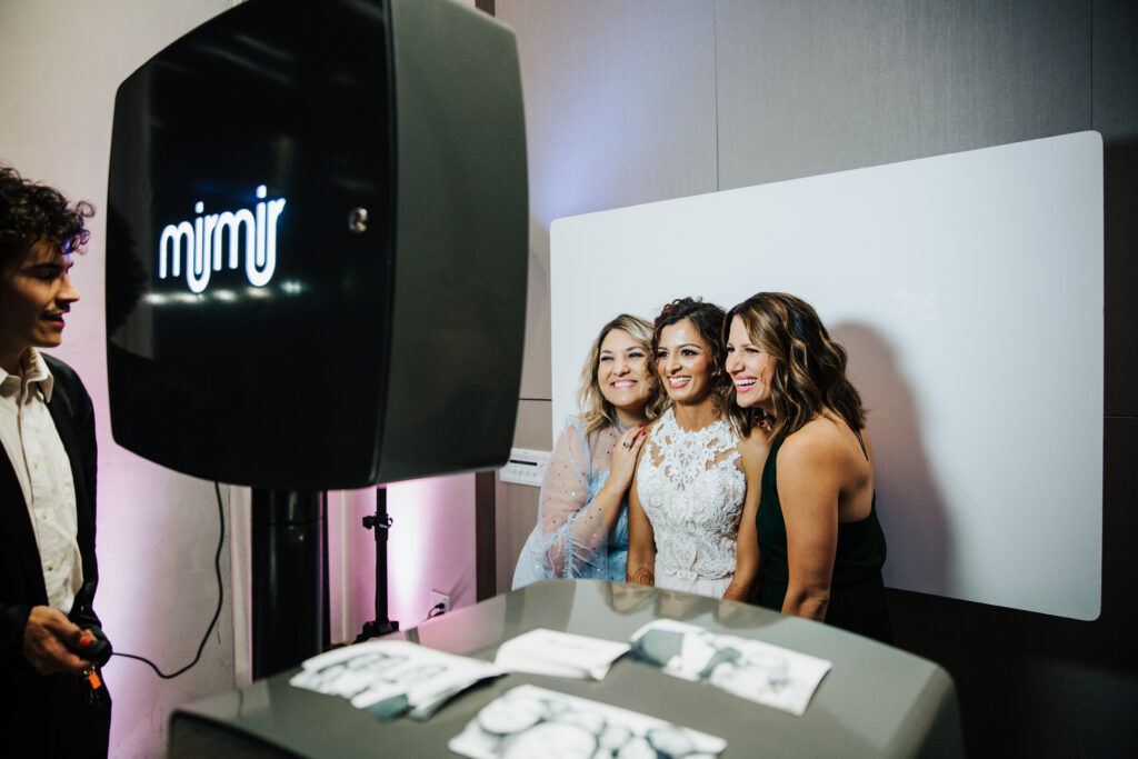 Three people pose closely in a photobooth, smiling at the camera. A man stands to the side, and printed photo strips are visible on a nearby surface.
