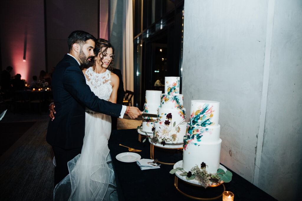 A couple in formal attire cuts a three-tiered white wedding cake with floral designs, standing on a table in a dimly lit venue.