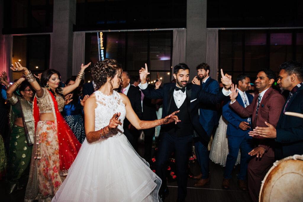 A bride and groom dance energetically among a group of people in colorful attire at a wedding reception.