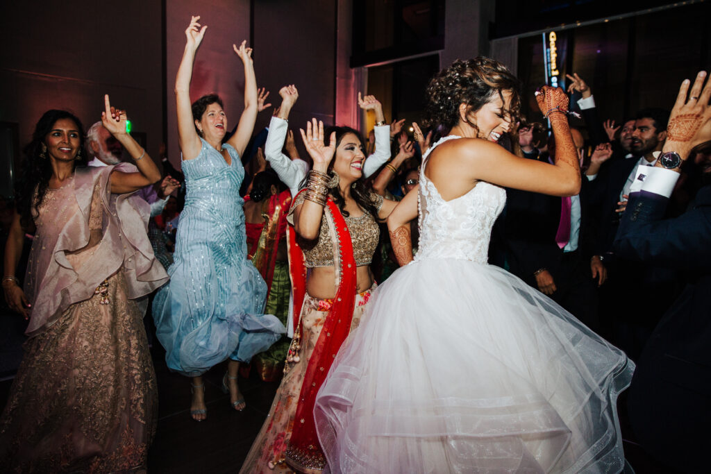 A group of people in formal attire, including a bride in a white dress, dance energetically at a party.