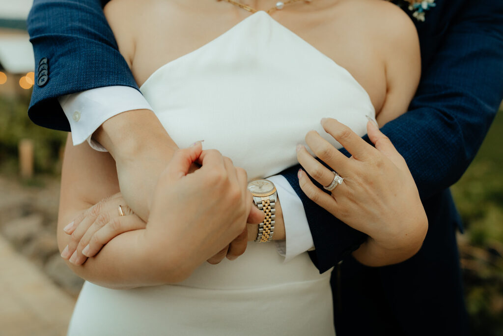 A couple in formal attire embraces; the woman in a white dress, and the man in a navy suit, showcasing a wristwatch and ring.