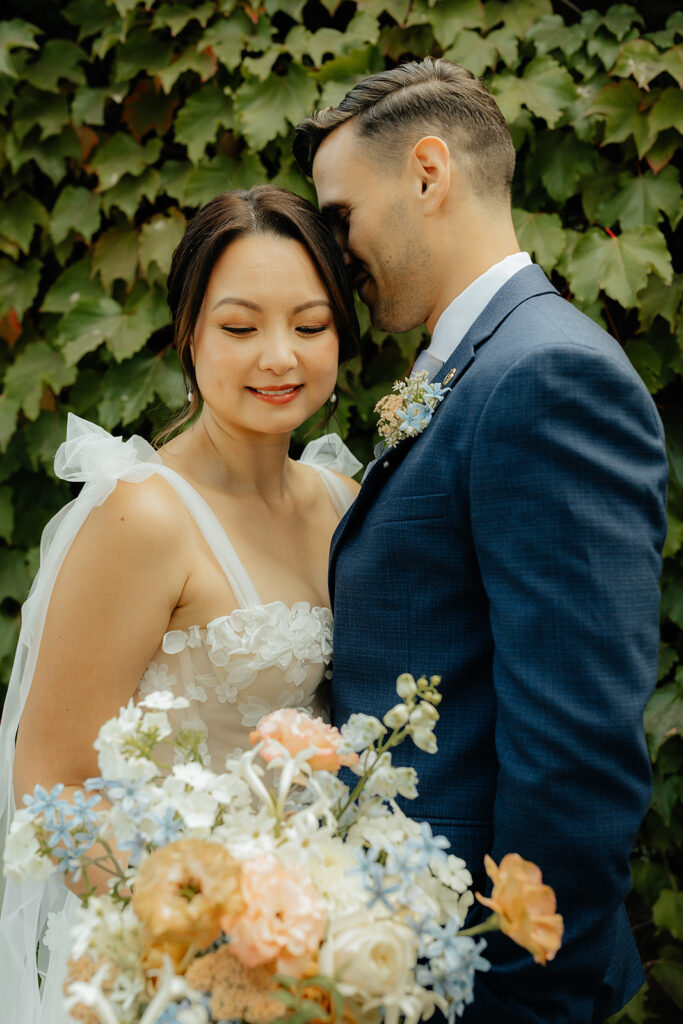 A couple in wedding attire stands close, smiling softly. The bride holds a bouquet of flowers. They are in front of a leafy green backdrop.