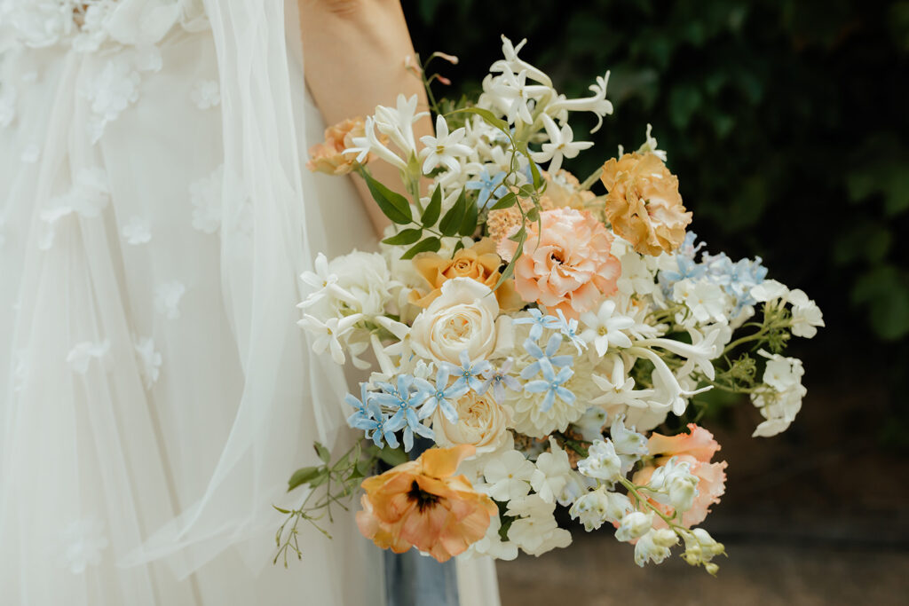 A bride holding a bouquet with white, peach, and blue flowers. The bouquet includes roses and other mixed blooms, set against a dark green leafy background.