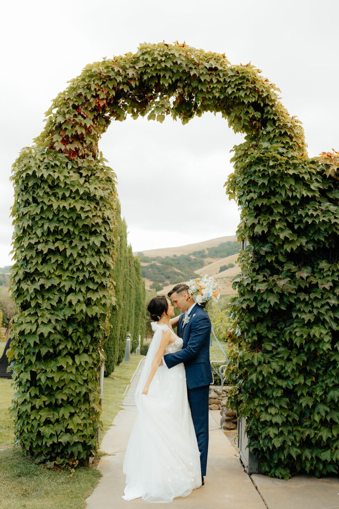 A couple in wedding attire embraces under an ivy-covered archway, set against a backdrop of trees and hills.