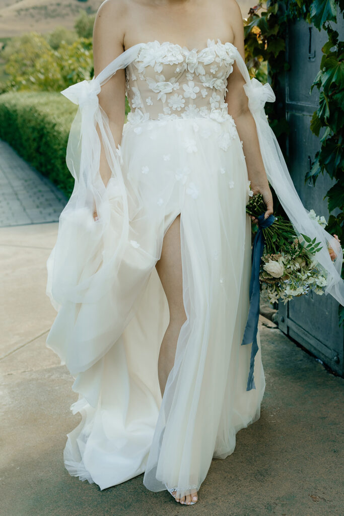 A person in an off-shoulder, floral appliqué wedding dress holds a bouquet while walking. The dress features a thigh-high slit.