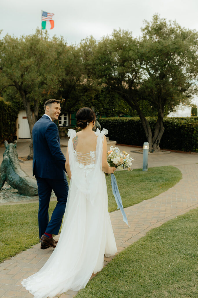 Bride and groom walk on a stone path in a garden setting, with trees and flags visible in the background. The bride holds a bouquet.