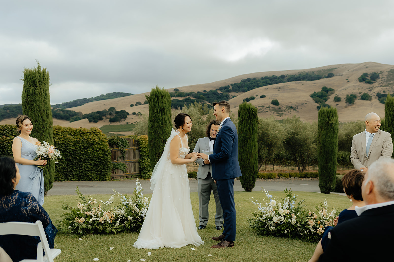 A couple stands at an outdoor wedding ceremony, exchanging vows in front of a small gathering. The setting features greenery and rolling hills in the background.