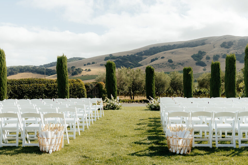Outdoor wedding ceremony setup with rows of white chairs facing an altar, set against rolling hills, trees, and a cloudy sky.