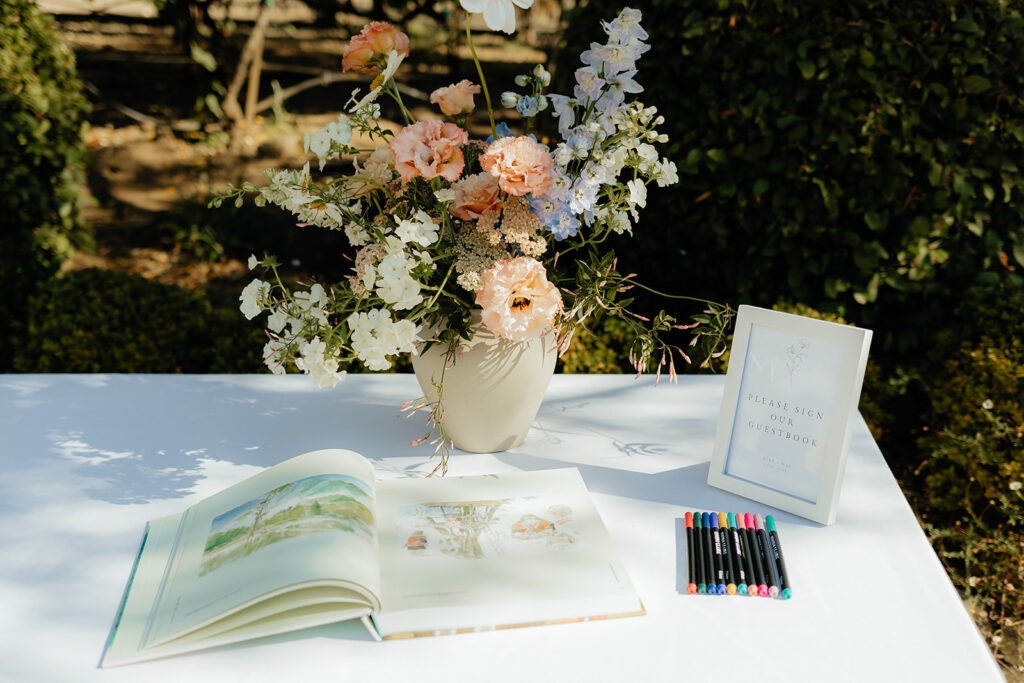 A table outdoors with an open book, a set of colored pens, a vase of pastel flowers, and a sign requesting guests to sign a guestbook.