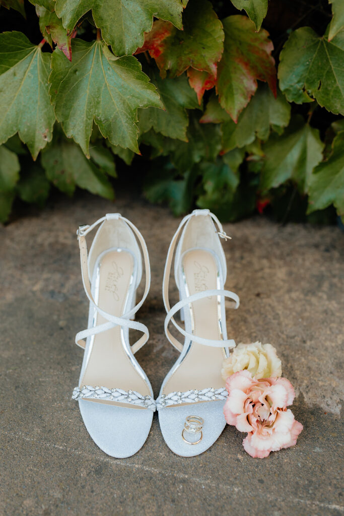 White strappy high heels placed on the ground, with decorative details on the front strap. A small bouquet of pink and yellow flowers is next to the shoes. Green leaves are in the background.