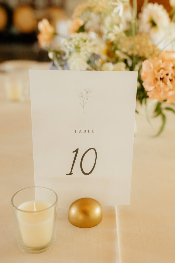 Table sign with the number 10, placed on a light-colored tablecloth. A small candle and a golden orb are nearby. Floral centerpiece visible in the background.