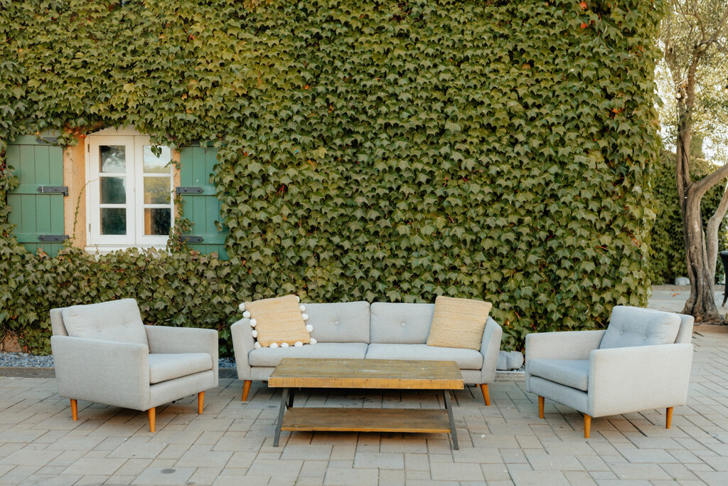 Outdoor patio set with a gray sofa, two armchairs, and a wooden coffee table on a paved area. Background features a green ivy-covered wall with white-framed window and green shutters.