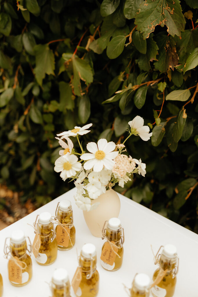 Small glass bottles with corks are displayed on a white surface. Each bottle contains a golden liquid and has a tag reading "Limoncello." A small vase holds white flowers in the background.