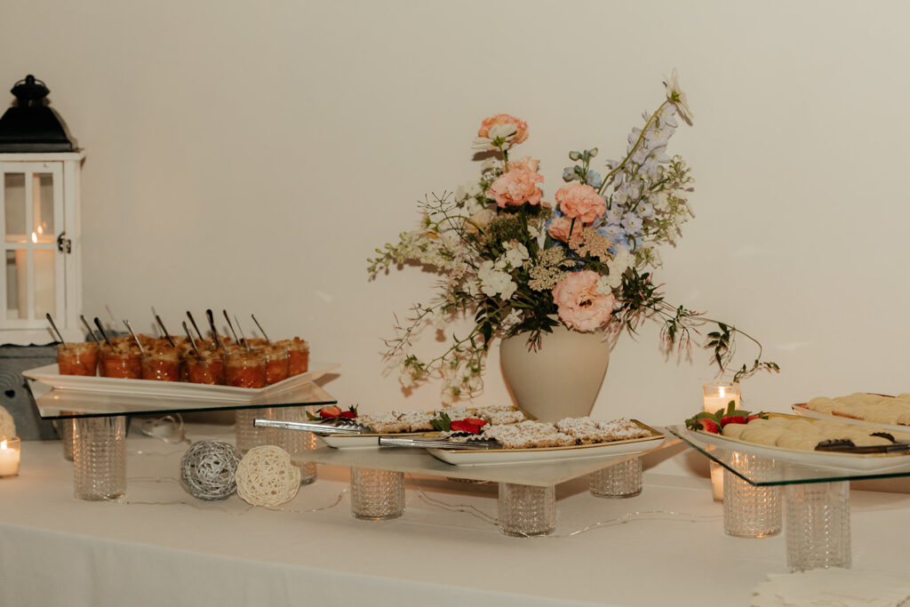 A table displaying a floral arrangement, trays of assorted pastries, desserts in cups with spoons, two decorative balls, and candles, set against a plain wall.
