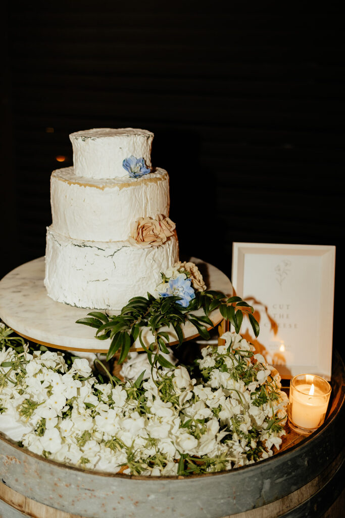 Three-tier white cake with textured frosting, adorned with blue and beige flowers. It's on a wooden barrel surrounded by white flowers, next to a lit candle and framed sign.