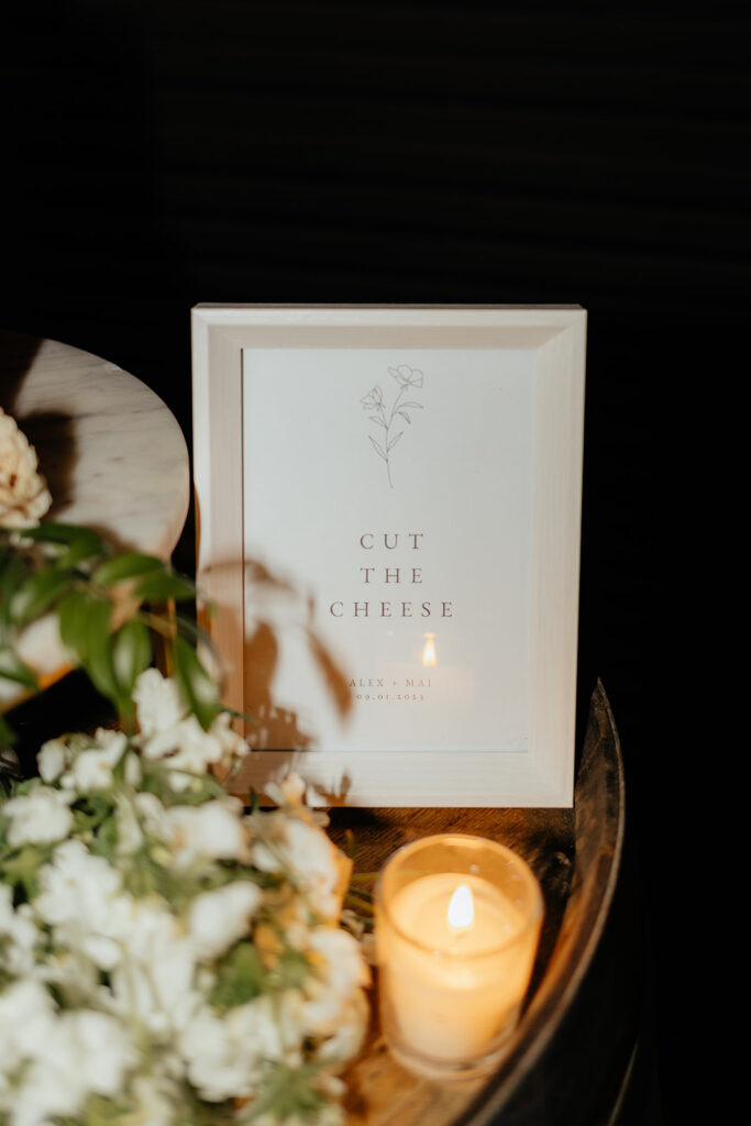 Framed sign with "Cut the Cheese" text beside a lit candle and white flowers on a dark surface.