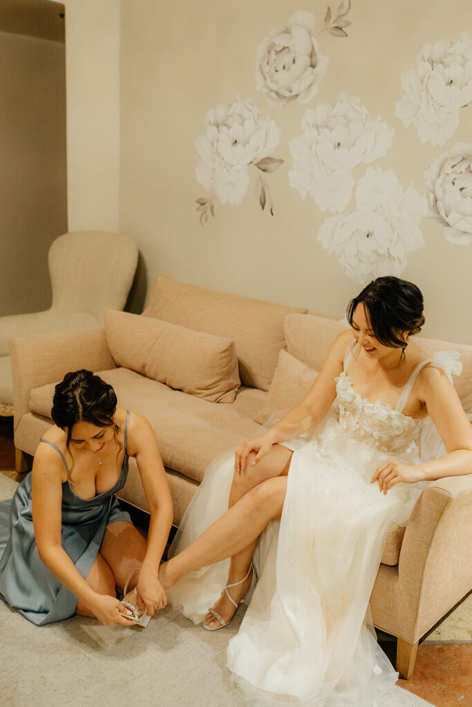 A woman in a wedding dress sits on a couch as another woman in a blue dress helps her with her shoe. They are in a room with floral wall decor.